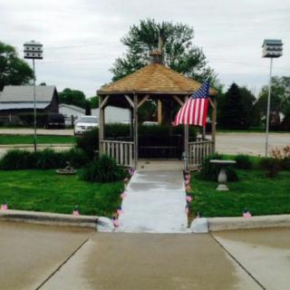 A gazebo with an American flag flanked by birdhouses stands at the end of a paved walkway, surrounded by green grass and shrubs under an overcast sky.