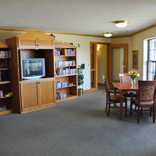 A cozy living room featuring a wooden bookshelf with a television, a round dining table with chairs, and ample natural light.