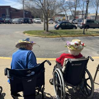 Two elderly people in wheelchairs wearing decorative hats sit outside near a parking lot on a sunny day.