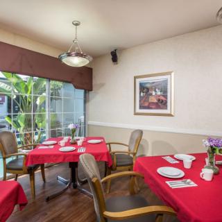 A cozy dining room with tables covered in red tablecloths, set for a meal, featuring comfortable chairs and a bright window view.