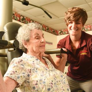 An elderly woman receives assistance from a healthcare worker while using gym equipment.