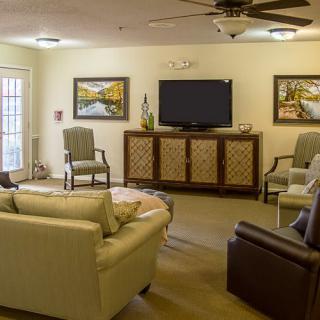 A cozy living room with a sofa, chairs, and a television, decorated with paintings and a ceiling fan.