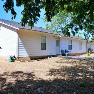 A sunny day view of a one-story house with a yard and chairs under the shade of trees.
