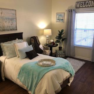 A cozy bedroom with a neatly made bed adorned with soft pillows and a turquoise throw blanket. A bedside table holds a lamp, while a welcome sign hangs above the window.