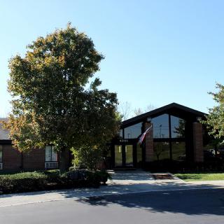 A building with large windows surrounded by trees and a parking lot under a clear blue sky.