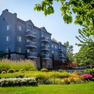 A view of a residential building surrounded by a beautifully landscaped garden with colorful flowers and lush trees on a sunny day.