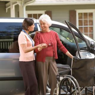 A caregiver assisting an elderly woman with a wheelchair next to a car.