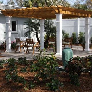 A serene garden patio with a wooden pergola and outdoor seating surrounded by lush plants.
