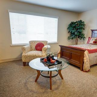 A cozy bedroom featuring an armchair, a glass coffee table, and a bed, with natural light streaming through the window.