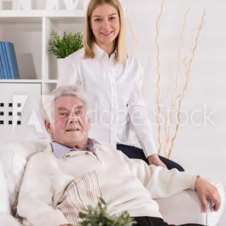 An elderly man sitting comfortably in a chair with a young woman standing beside him, both smiling in a cozy home setting.