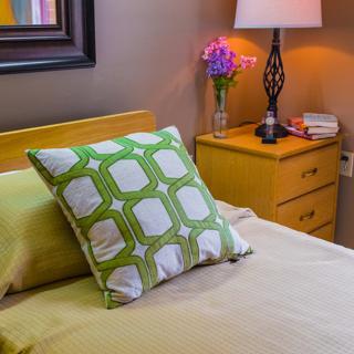 A cozy bedroom with a patterned pillow on the bed and a bedside table featuring a lamp and flowers.