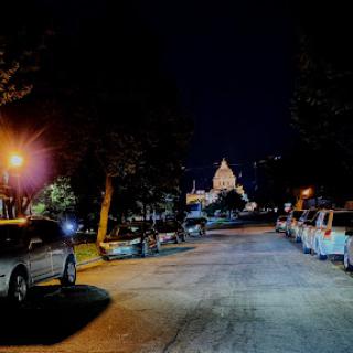 A quiet street scene at night, illuminated by streetlights and lined with parked cars.