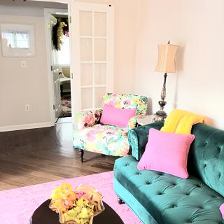 A bright and colorful living room featuring a teal sofa, a floral armchair, and vibrant pink pillows.