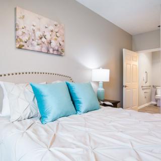 A cozy bedroom with a neatly made bed featuring white linens and blue accent pillows, complemented by a bedside lamp and wall art.