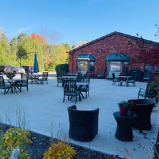 A spacious outdoor patio with tables and chairs, set against a brick building and surrounded by greenery under a clear sky.
