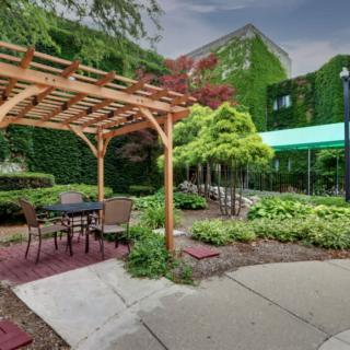 A tranquil garden scene with a pergola and outdoor seating area surrounded by lush greenery and an ivy-covered building.