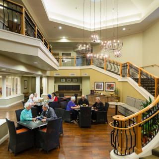 A spacious interior lounge with people gathered around tables, featuring a grand staircase and chandelier.