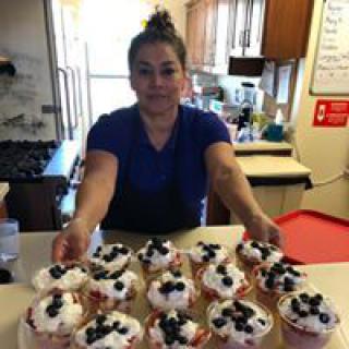 A woman presenting a batch of freshly made cupcakes topped with blueberries in a kitchen.