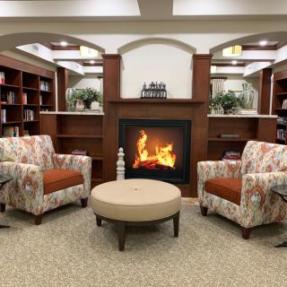 A cozy living room with two patterned chairs facing a warm fireplace, surrounded by bookshelves.