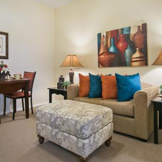A cozy living room with a beige sofa adorned with colorful pillows, complemented by an ottoman. The space is decorated with modern artwork and ambient lighting from table lamps.