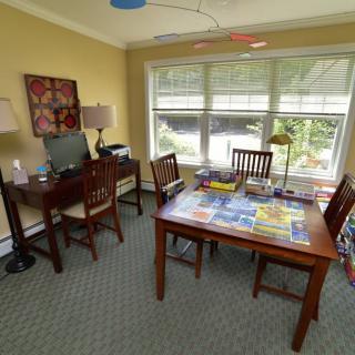 A cozy home office with a desk, chair, and natural light streaming through a large window. A puzzle is laid out on the table.