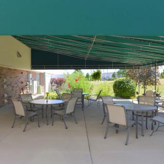 An outdoor patio area with tables and chairs, shaded by a green awning, overlooking a garden on a sunny day.