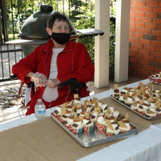 A woman wearing a mask sits at an outdoor table surrounded by delicious cupcakes.