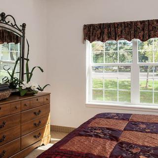 A cozy bedroom with a wooden dresser, mirror, and a bed covered with a colorful quilt next to a sunlit window.