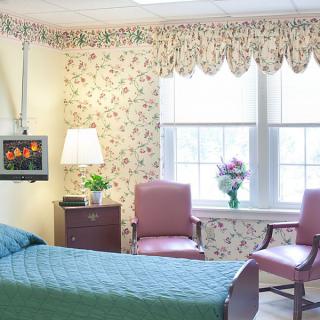 A cozy hospital room with a single bed, floral wallpaper, and two chairs by the window.