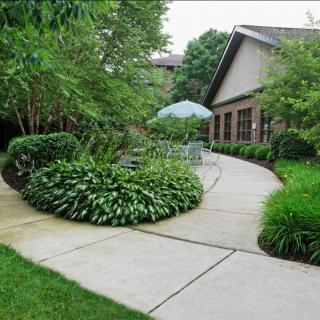 A tranquil garden with a winding concrete pathway lined with lush greenery and a cozy patio area under an umbrella.