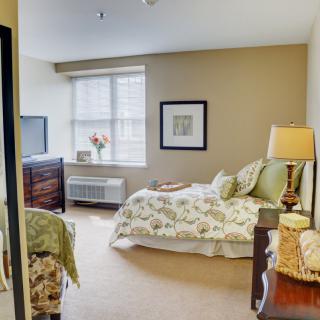 A cozy bedroom with a neatly made bed, bedside lamp, and natural light coming through the window.