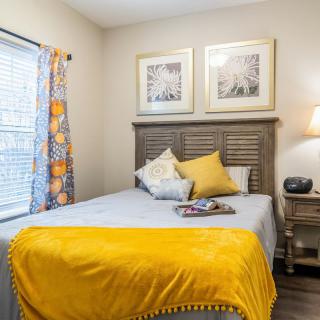 A cozy bedroom featuring a bed with a yellow blanket and decorative pillows, complemented by artwork on the wall and a lamp on the nightstand.