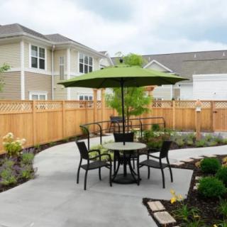 A cozy patio setup with a round table, chairs, and a green umbrella, surrounded by landscaped garden and wooden fencing.