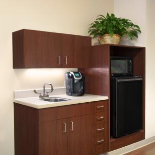 A compact kitchenette featuring a coffee maker, sink, cabinets, microwave, mini fridge, and a potted plant.
