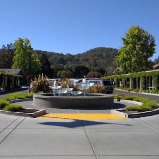 A scenic view featuring a central fountain surrounded by greenery, with mountains in the background and a clear blue sky above.
