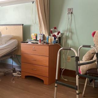 A hospital room with a bed, a dresser with personal items, and a walker with a teddy bear resting on it.