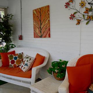 A cozy living room featuring a white wicker sofa with orange cushions, colorful patterned pillows, and decorative wall art.