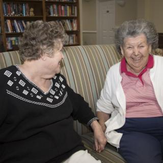 Two elderly women sit together on a couch, smiling and enjoying each other's company in a library setting.