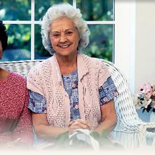 An elderly woman smiling while seated indoors with a view of the garden through the window.