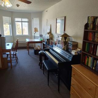 A cozy living room with a piano, bookshelf, and workspace by the window.