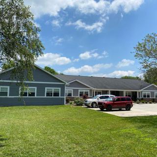 A suburban house with a well-maintained lawn, two cars parked in the driveway under a blue sky with scattered clouds.