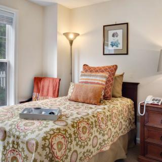 A cozy bedroom with a neatly made bed, decorative pillows, and a standing lamp beside a window.