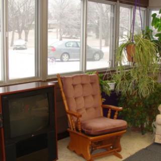 A cozy indoor space with a glider chair, television, and various plants near large windows.