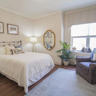 A cozy bedroom with a neatly made bed, a standing lamp, a decorative mirror, and a comfortable chair by the window.