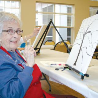 An elderly woman participates in a painting class, working on a canvas with an outline of a tree.