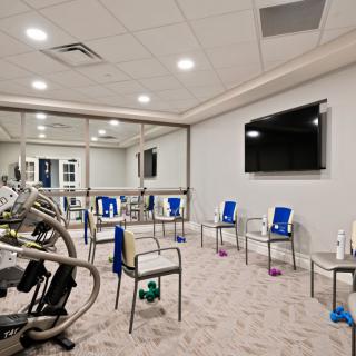 A well-lit exercise room with chairs arranged in a circle, featuring a large television and mirrored walls.