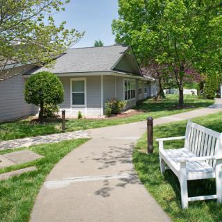 A peaceful suburban scene with a white bench along a pathway, surrounded by trees and a small house.