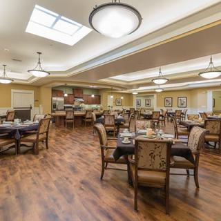 A spacious and elegantly designed dining area with neatly arranged tables and chairs, featuring warm lighting and a wooden floor.
