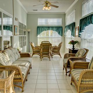 A bright sunroom featuring wicker furniture and a dining area, with large windows and a ceiling fan.