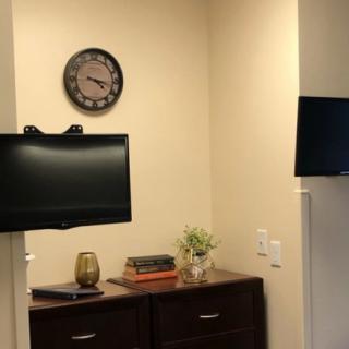 A small office corner with two televisions, a clock on the wall, and a cabinet topped with books and a plant.